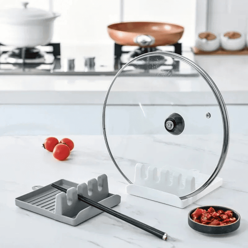 Suporte e descanso para tampas e espátulas cinza com 2 hashis em cima, e suporte com tampa de vidro, ao lado de tomates e aperitivos