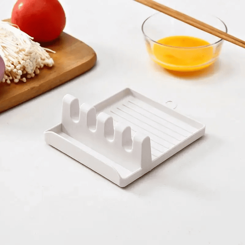 Suporte e descanso para tampas e espátulas branco sobre mesa branca, com copo de molho amarelo e bandeja de madeira com legumes